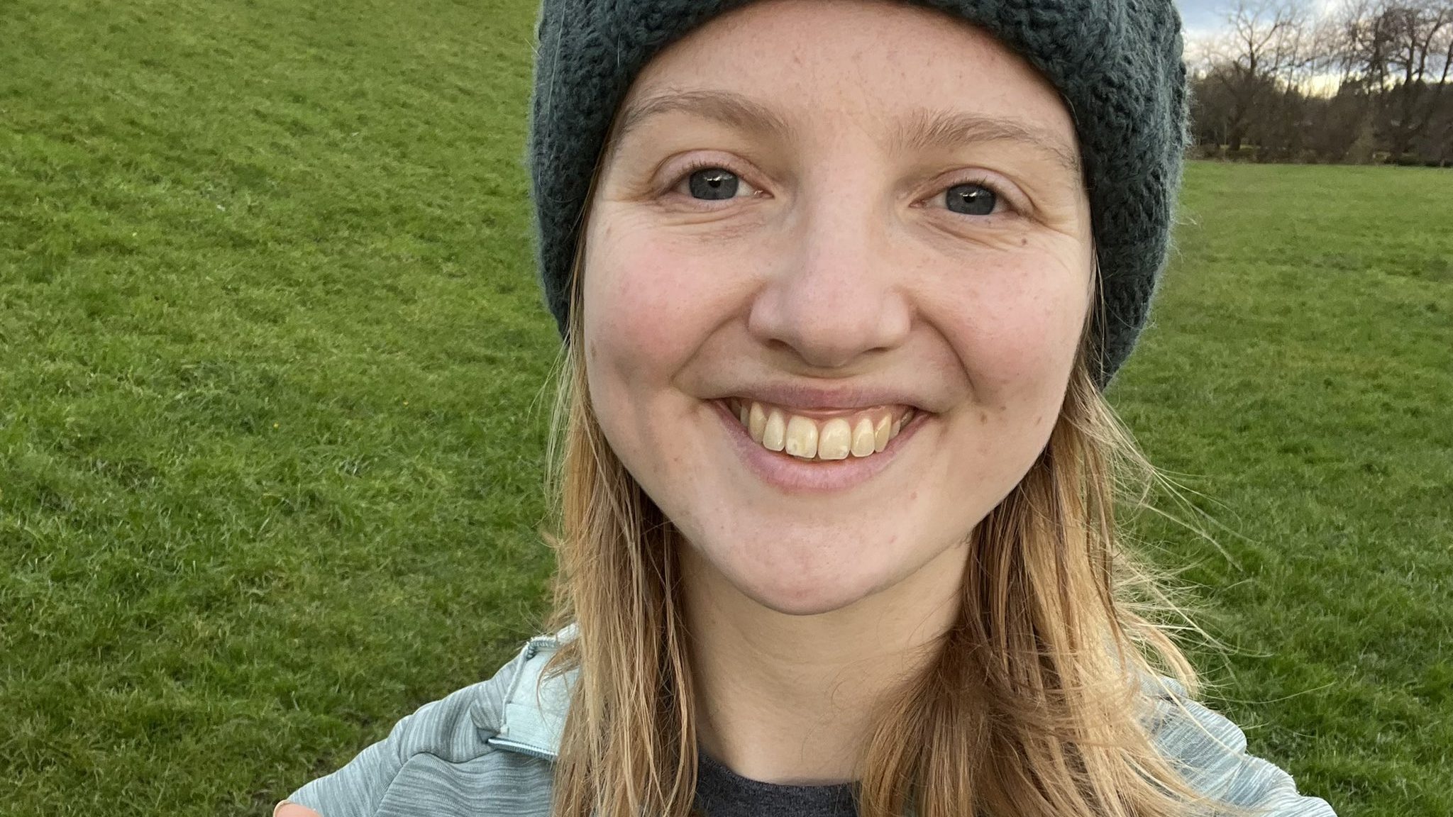 Charlie, a woman with dark blonde hair and blue eyes, is wearing a blue hat and blue fleece and is smiling. She has grass behind her. 